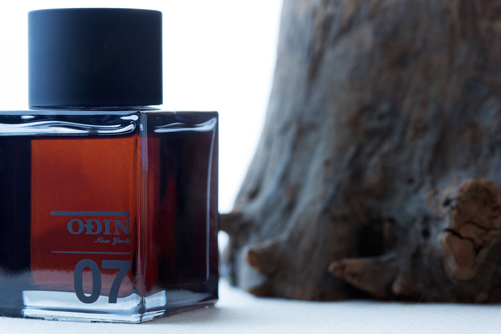 Image shows a glass bottle of Odin 07 Tanoke fragrance with a black cap.  It is positioned next to a blurred wood trunk.