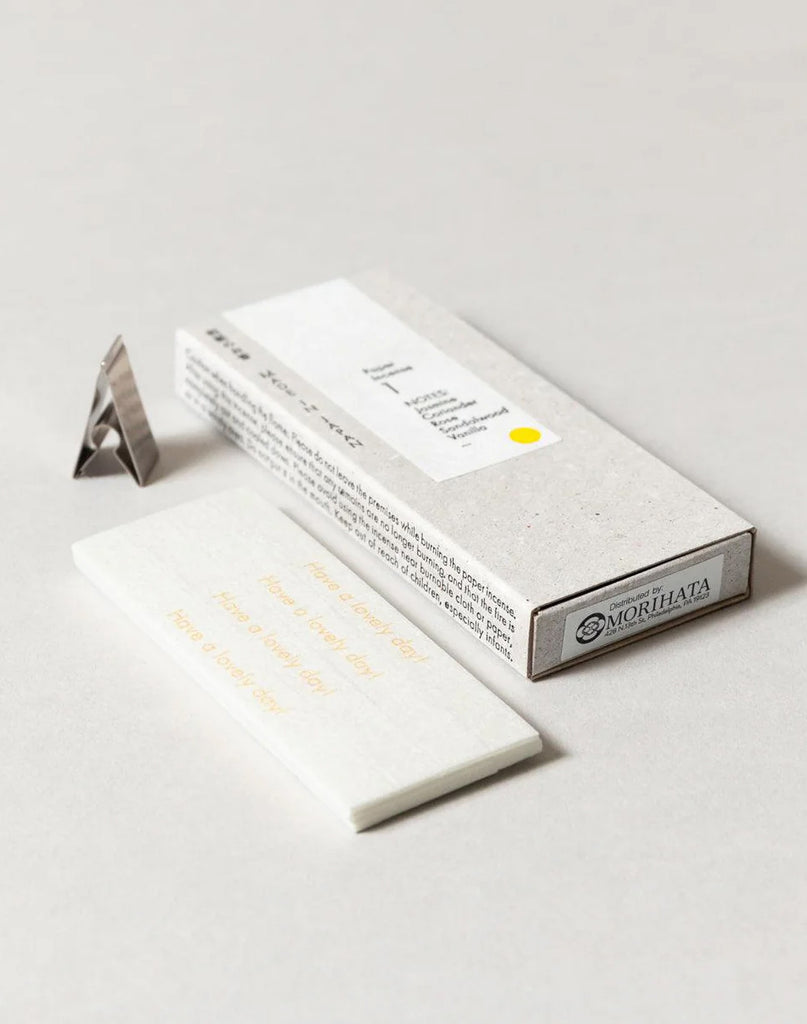2 of 3.  White paper incense stick packaging with text, metal incense holder and incense strips on a gray background