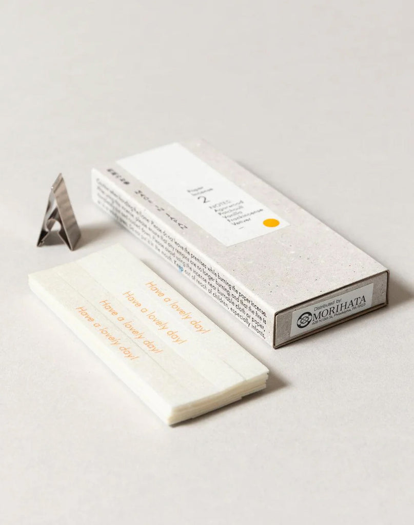 2 of 4.  White paper incense stick packaging with text, metal incense holder and incense strips on a gray background