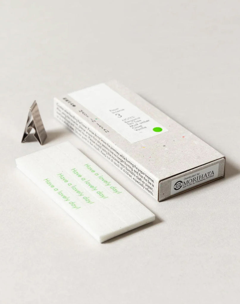 2 of 4.  White paper incense stick packaging with text, metal incense holder and incense strips on a gray background. Strips have text in green "Have a lovely day!"