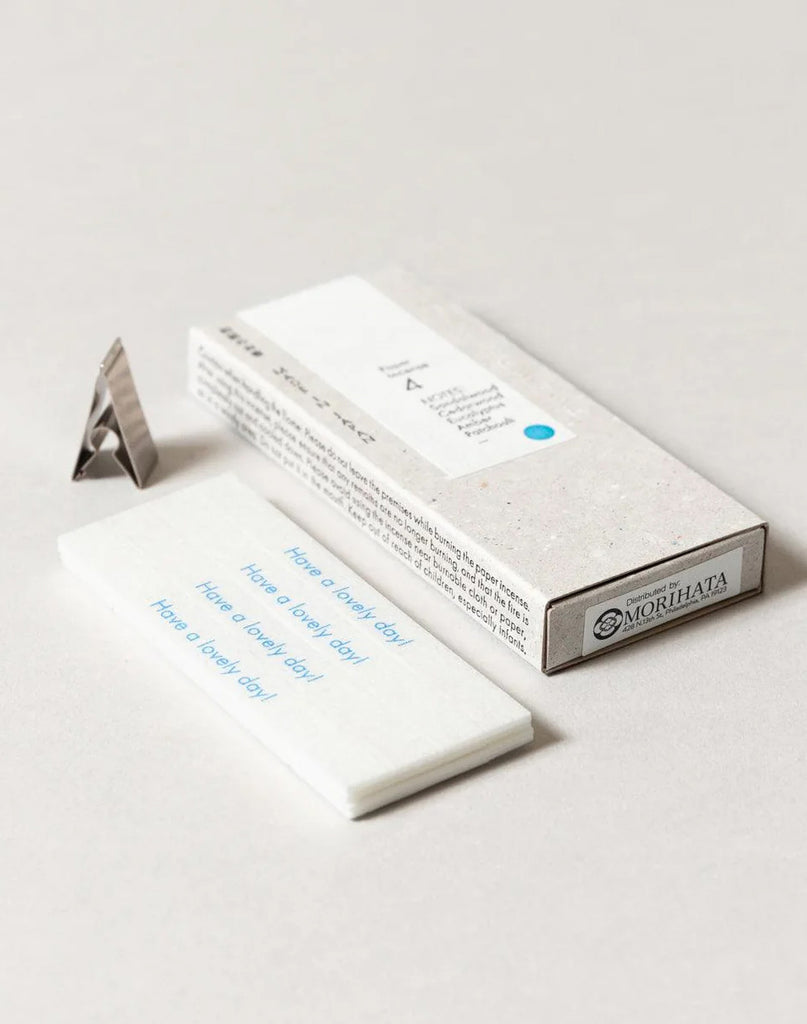 2 of 5.  White paper incense stick packaging with text, metal incense holder and incense strips on a gray background