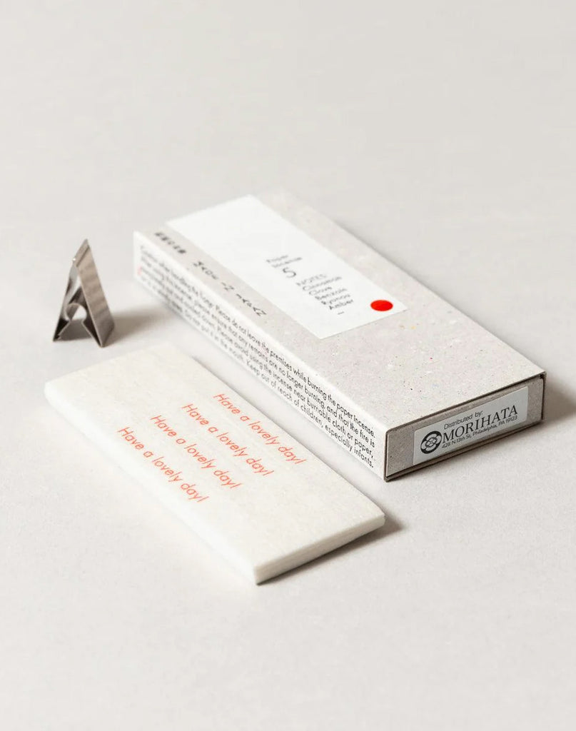 2 of 5.  White paper incense stick packaging with text, metal incense holder and incense strips on a gray background. Strips have text in red reading "Have a lovely day!"