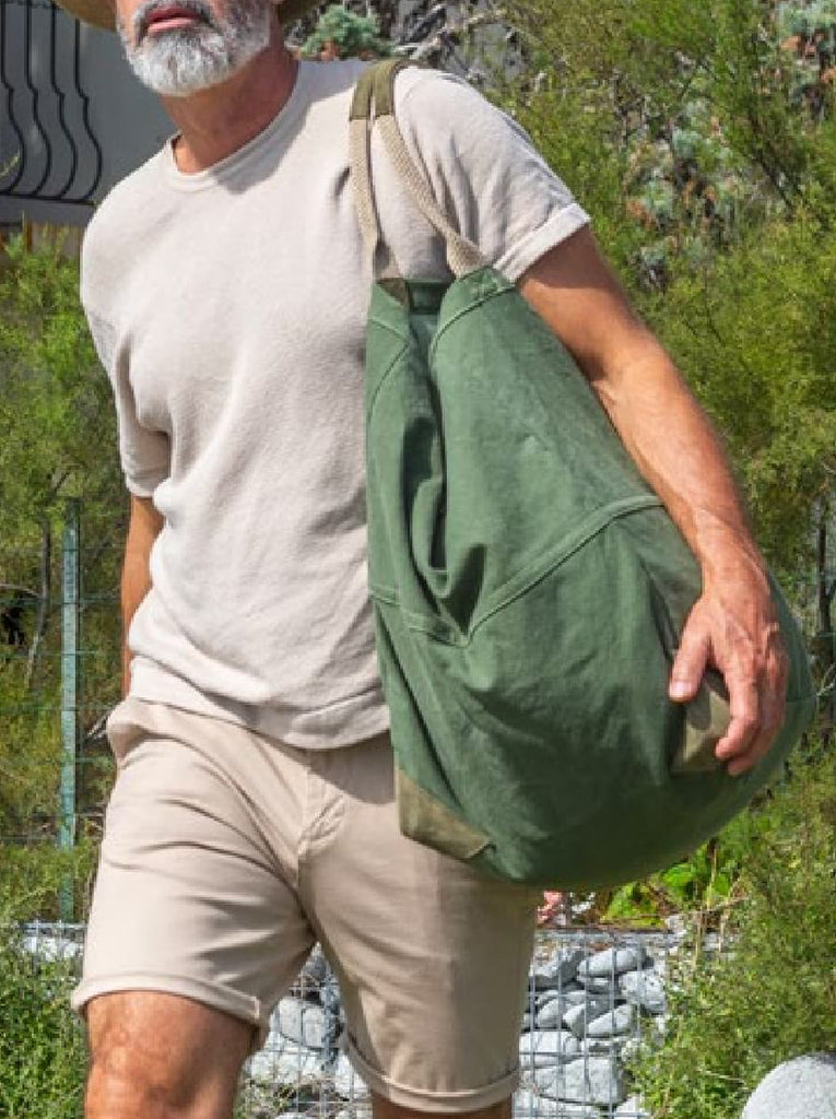 Man with white beard and tan beige t-shirt and shorts has the large tote in green around his shoulder.  The bag is hanging on his shoulder by the two cotton handles.   Background shows trees and brush.   Slide 5 of 5