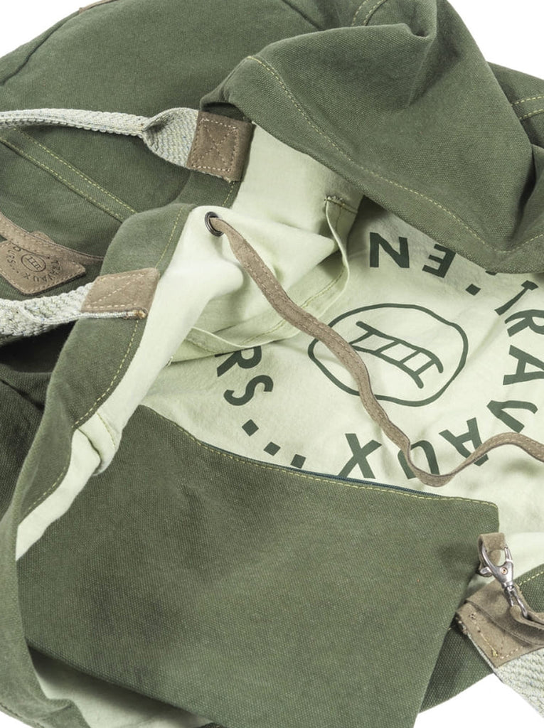 Partial Interior shot of green canvas bag with orange cotton interior.  Partial "Travaux" in green can be seen on the printed off white cotton interior.  Khaki contrast stitching at seems and hems.  Travaux En Cours logo patch on exterior of the bag in khaki green.  Slide 4 of 5