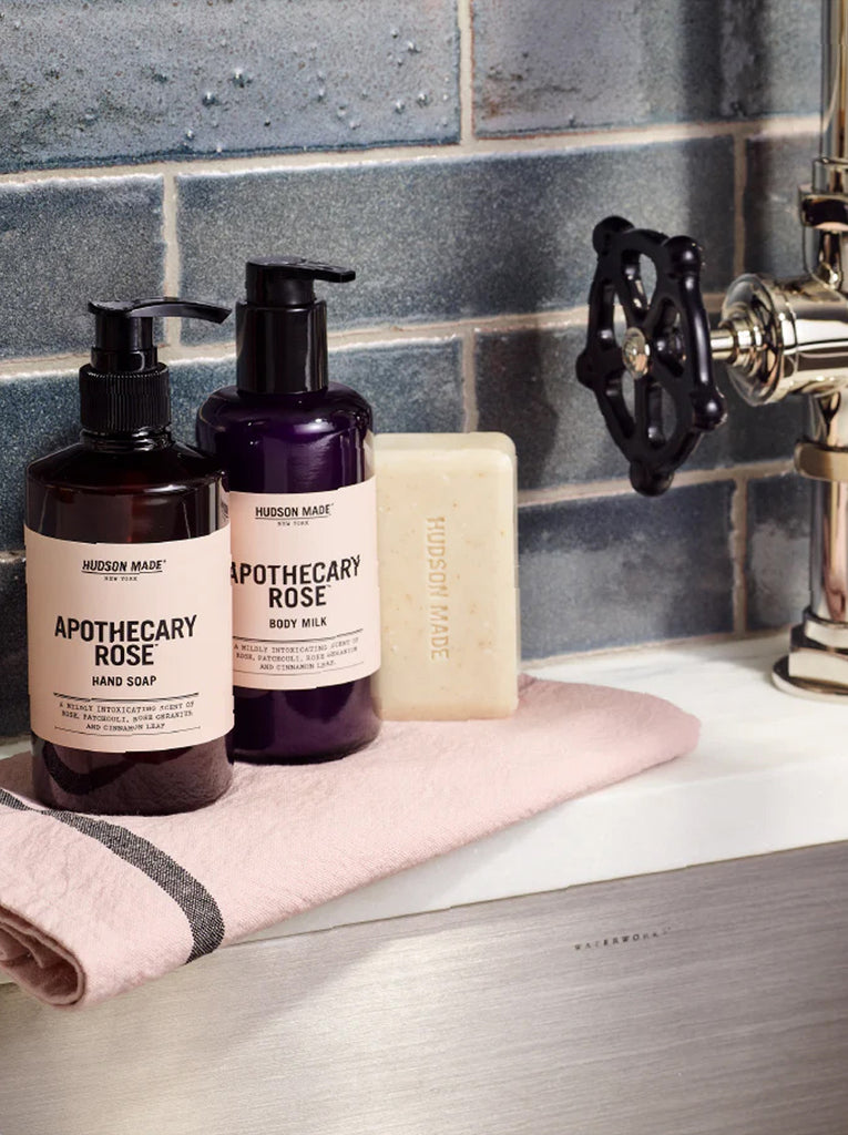 Two bottles by Hudson Made.  First pump bottle is Apothecary Rose hand soap and the second bottle is Apothecary Rose Body milk.  The bottles are on a sink counter with a facet in the background.  Slide 3 of 3