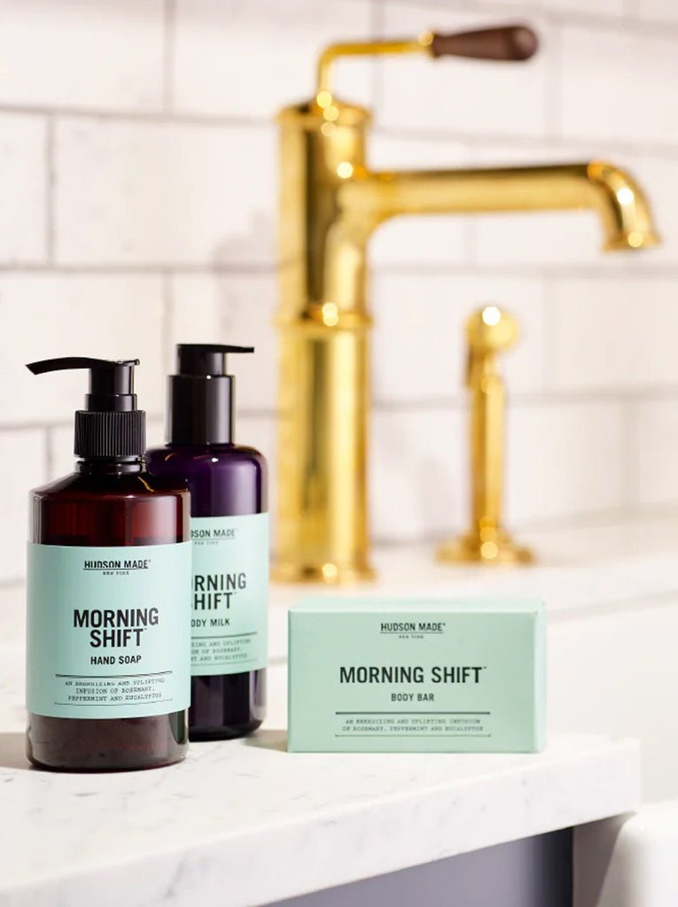 Two bottles by Hudson Made.  First pump bottle is Morning Shift hand soap and the second bottle is Morning Shift Body milk.  There is a Morning Shift boxed soap. next to the bottles.  The soaps are on a sink counter with a brass facet in the background.  Slide 3 of 4