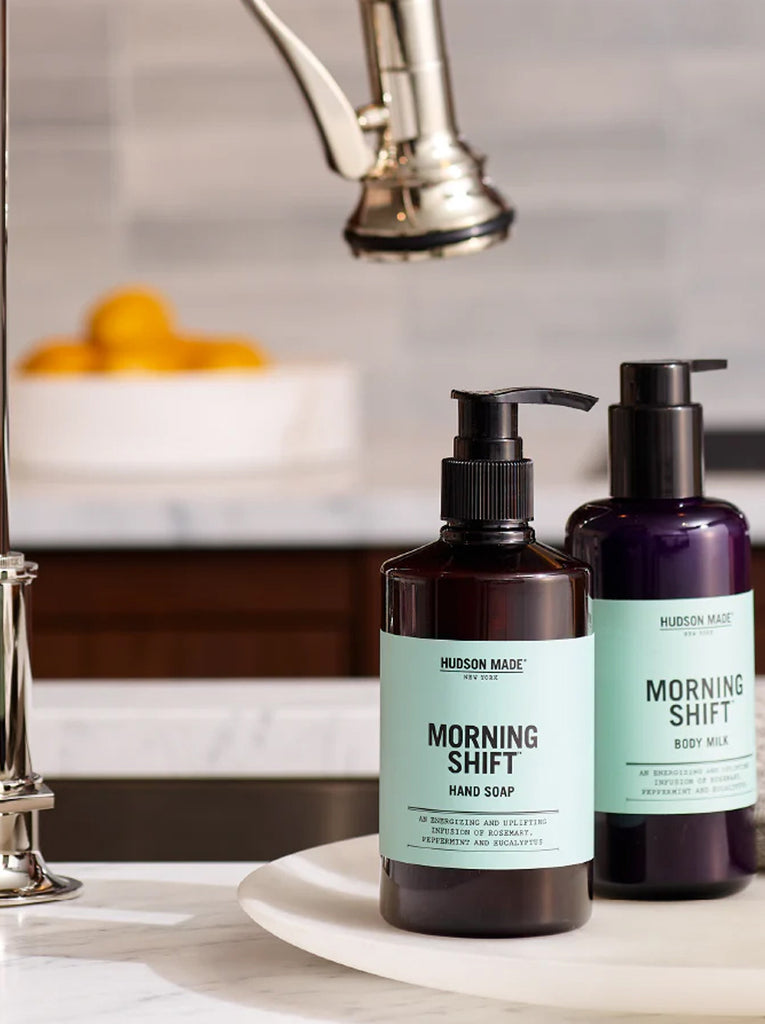 Two bottles by Hudson Made.  First pump bottle is Morning Shift hand soap and the second bottle is Morning Shift Body milk.    The soaps are on a sink counter with a chrome facet in the background.  Slide 3 of 4