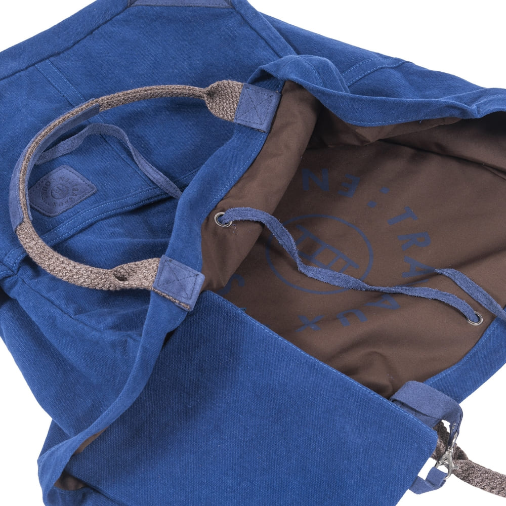 Partial Interior shot of indigo canvas bag with charcoal gray cotton interior.  Partial "Travaux" in blue print can be seen on the printed cotton.  Blue tonal contrast stitching at seems and hems.  Travaux En Cours logo patch on exterior of the bag.  Slide 4 of 4