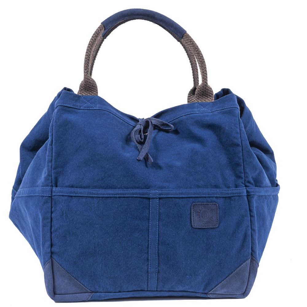 Front view of the indigo tote bag with two curved handles with leather trim. Indigo leather tie in the center of bag with indigo logo patch on center right of bag. Slide 2 of 4