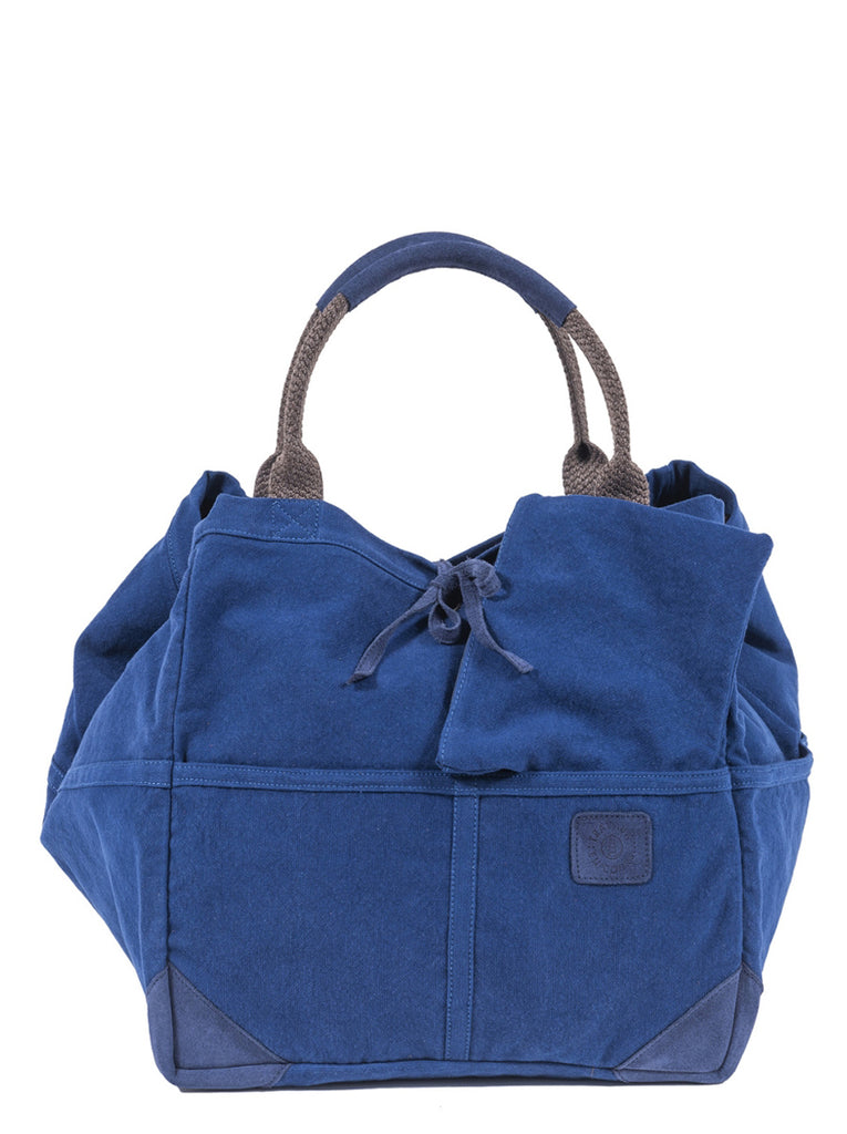 Front view of the indigo tote bag with two curved handles with leather trim. Indigo leather tie in the center of bag with indigo logo patch on center right of bag.  Indigo zip pouch is tucked in the exterior pocket and laying across as it can be removed.  Slide 1 of 4